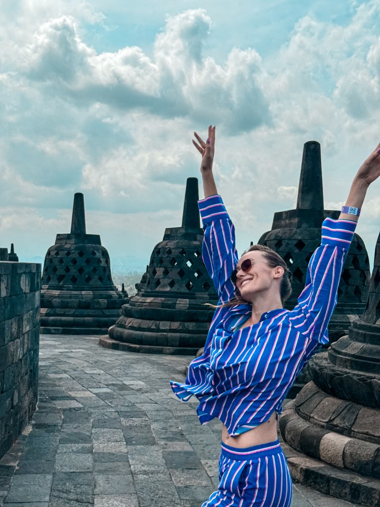 Carlijn smiling with raised arms between Borobudur stupas under a blue sky
