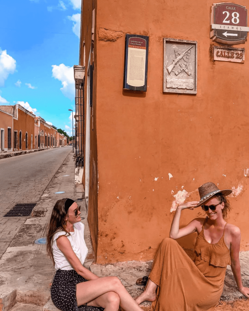 Two girls sitting on the corner of a street in Izamal Mexico, where all the buildings are yellow.