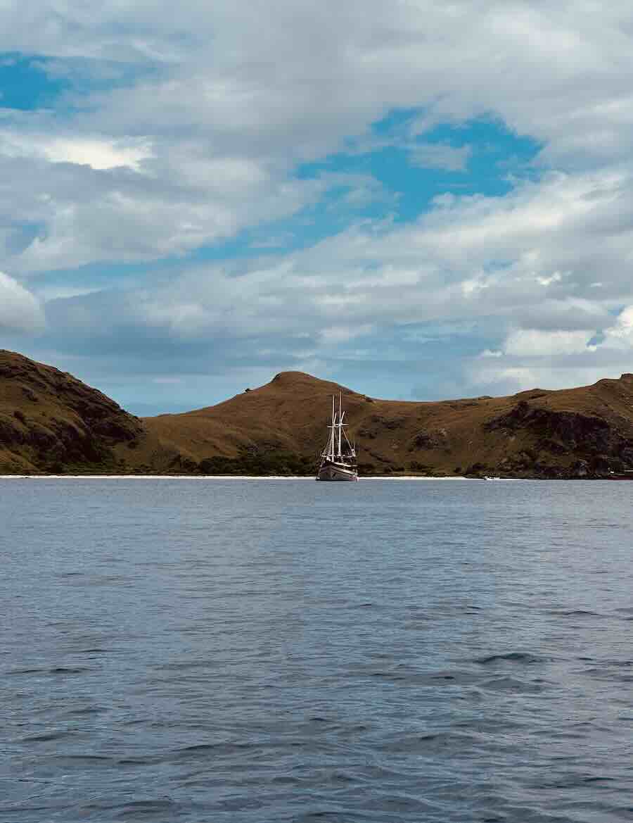 Phinisi liveaboard anchored in calm water between dry golden hills in Komodo National Park.