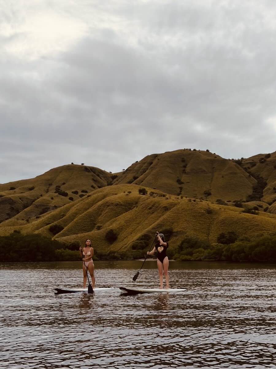 Two travelers paddleboarding on calm water beneath the dry golden hills of Komodo National Park.
