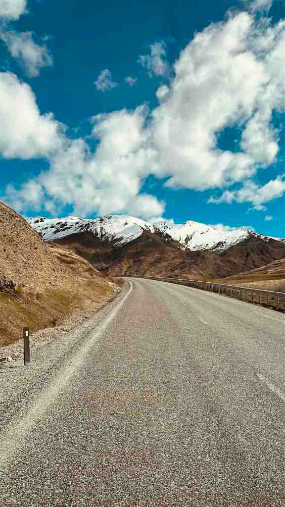 Open Crown Range Road leading toward snow-covered mountain peaks beneath a bright blue sky.