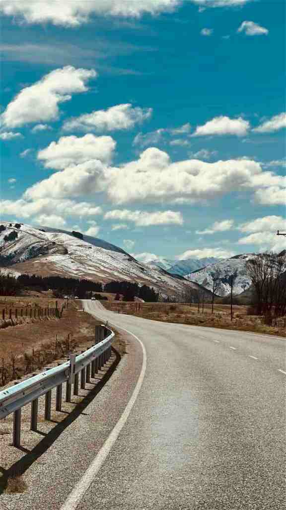 Mountain road with a guardrail beneath rugged snow-dusted peaks on the Lindis Pass drive.