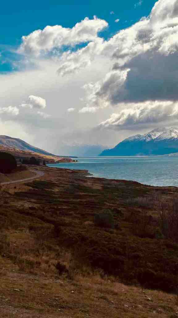 Lake Pūkaki and distant snow-covered mountains beneath dramatic clouds along Mount Cook Road.