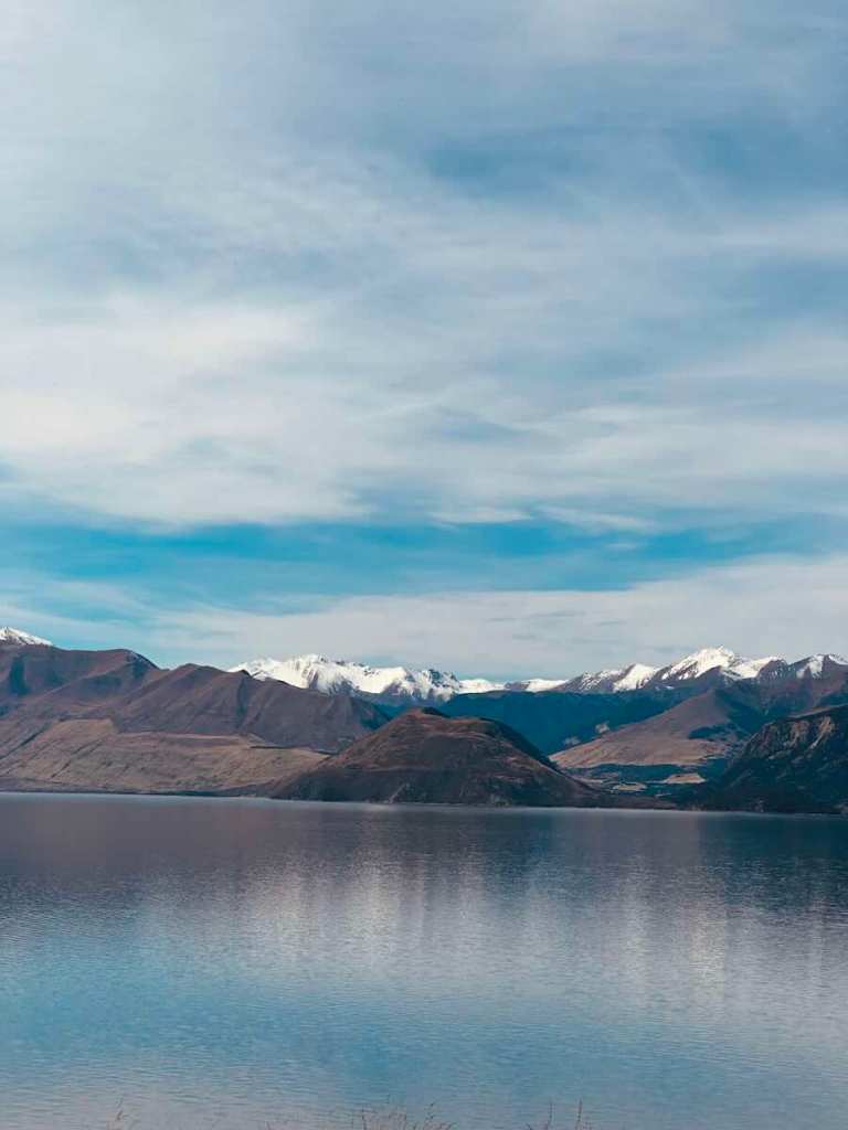 Calm Lake Hāwea surrounded by rugged mountains beneath an overcast sky.